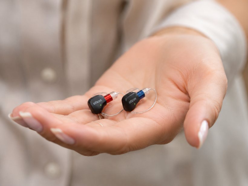 A pair of nearly invisible Oticon hearing aids resting in an open hand